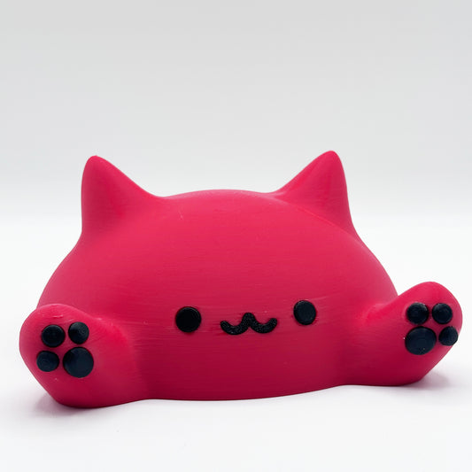 A small, red, cat-shaped desk toy with a rounded body, black eyes, and a cute black mouth resembling a squiggly line. It has two front paws extended forward, each with black paw pads. The toy appears to be made of a smooth, matte material. It's the Blob Cat Desk Decor from Highly Obtainable.