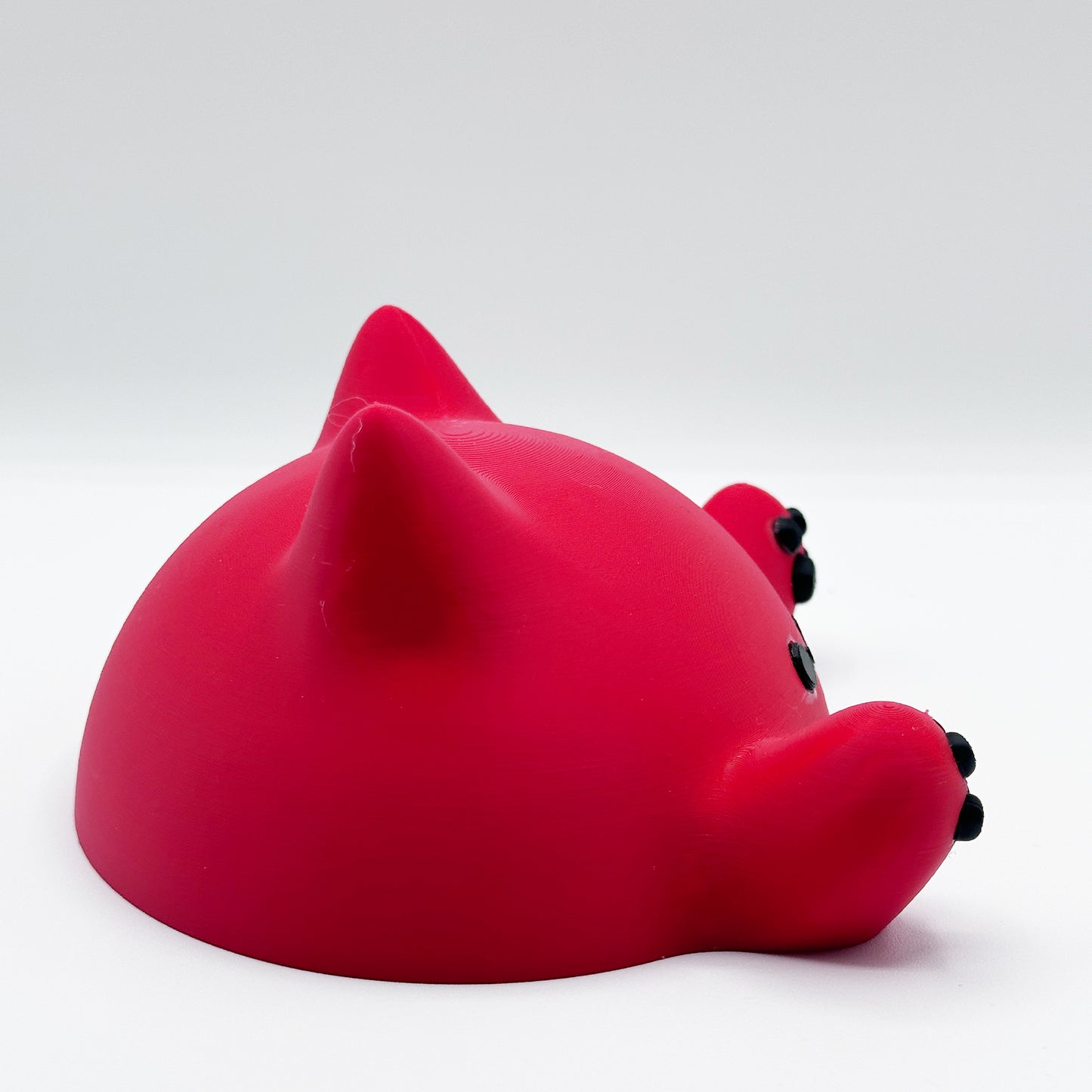 A small, round, red figurine resembling a cute creature with two pointed ears, black eyes, and small black paws placed in front. The Blob Cat Desk Decor by Highly Obtainable is positioned facing away at an angle, showing the back and side view against a plain white background.