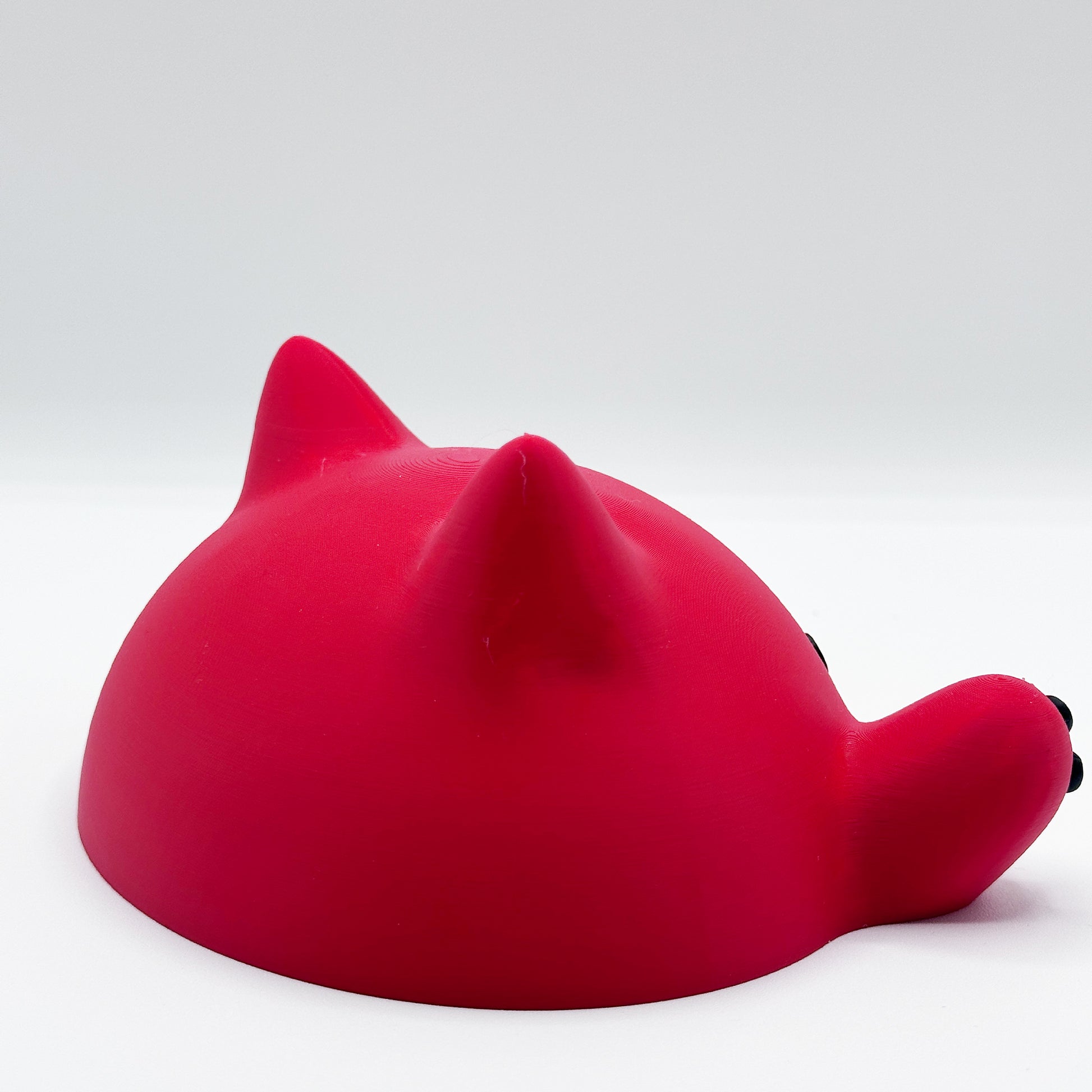 A bright red, dome-shaped object with two pointy ears and a small paw, resembling a cat, sits against a plain white background. This is the Blob Cat Desk Decor by Highly Obtainable.
