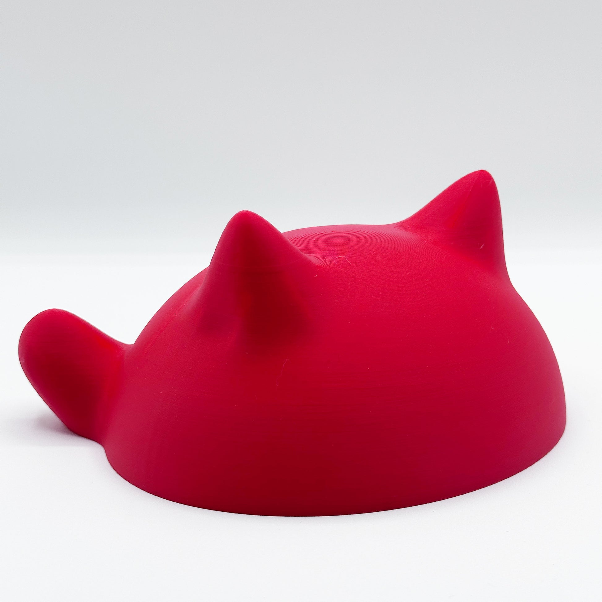 A bright red, dome-shaped object resembling a cat's head with ears, viewed from the back against a plain white background is the Blob Cat Desk Decor by Highly Obtainable.