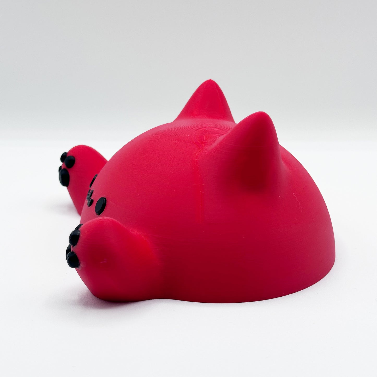 A red, plastic figure resembling a partially submerged cat, with visible ears, black eyes, and a black nose. The cat's left paw extends outward, showing black paw pads. The background is a plain, white surface features the Blob Cat Desk Decor by Highly Obtainable.