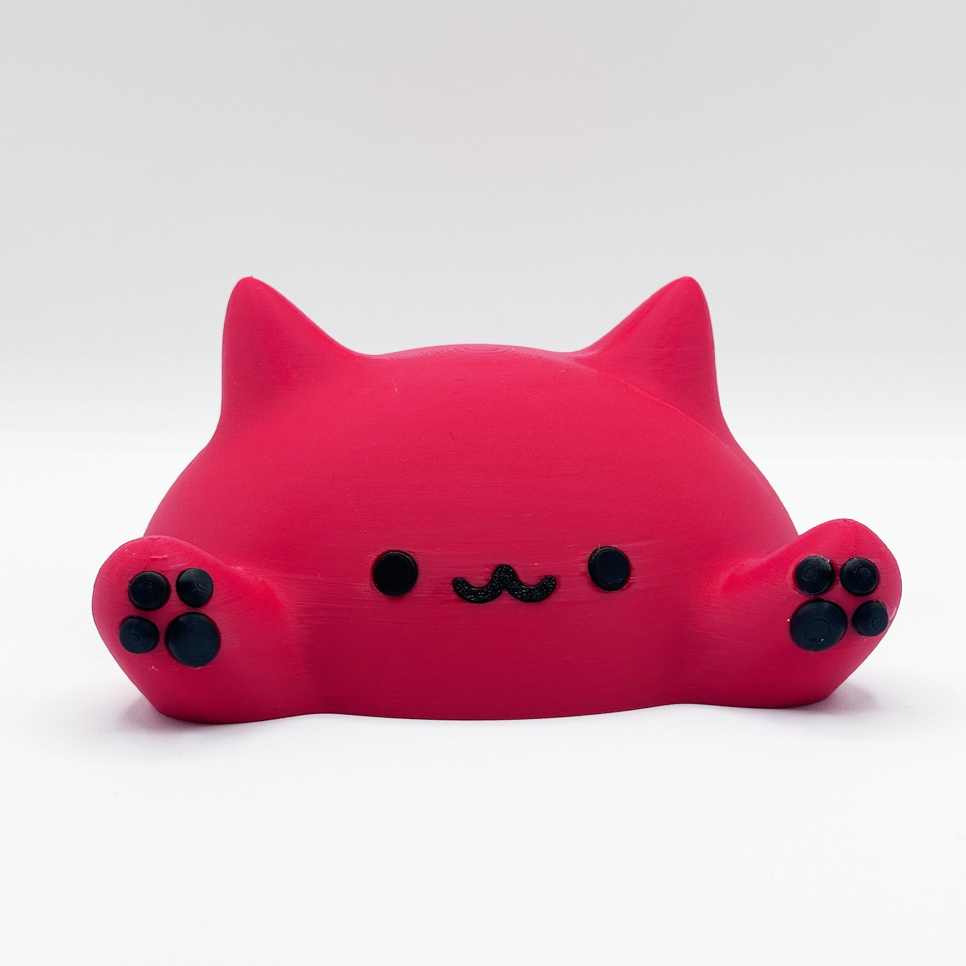 A small, bright pink, cat-like figure with a simple, cute face featuring black eyes, a tiny mustache-shaped mouth, and two upraised paws with black paw prints. The figure is designed in a minimalist, round style and is placed against a plain white background. This charming object is the Blob Cat Desk Decor by Highly Obtainable.