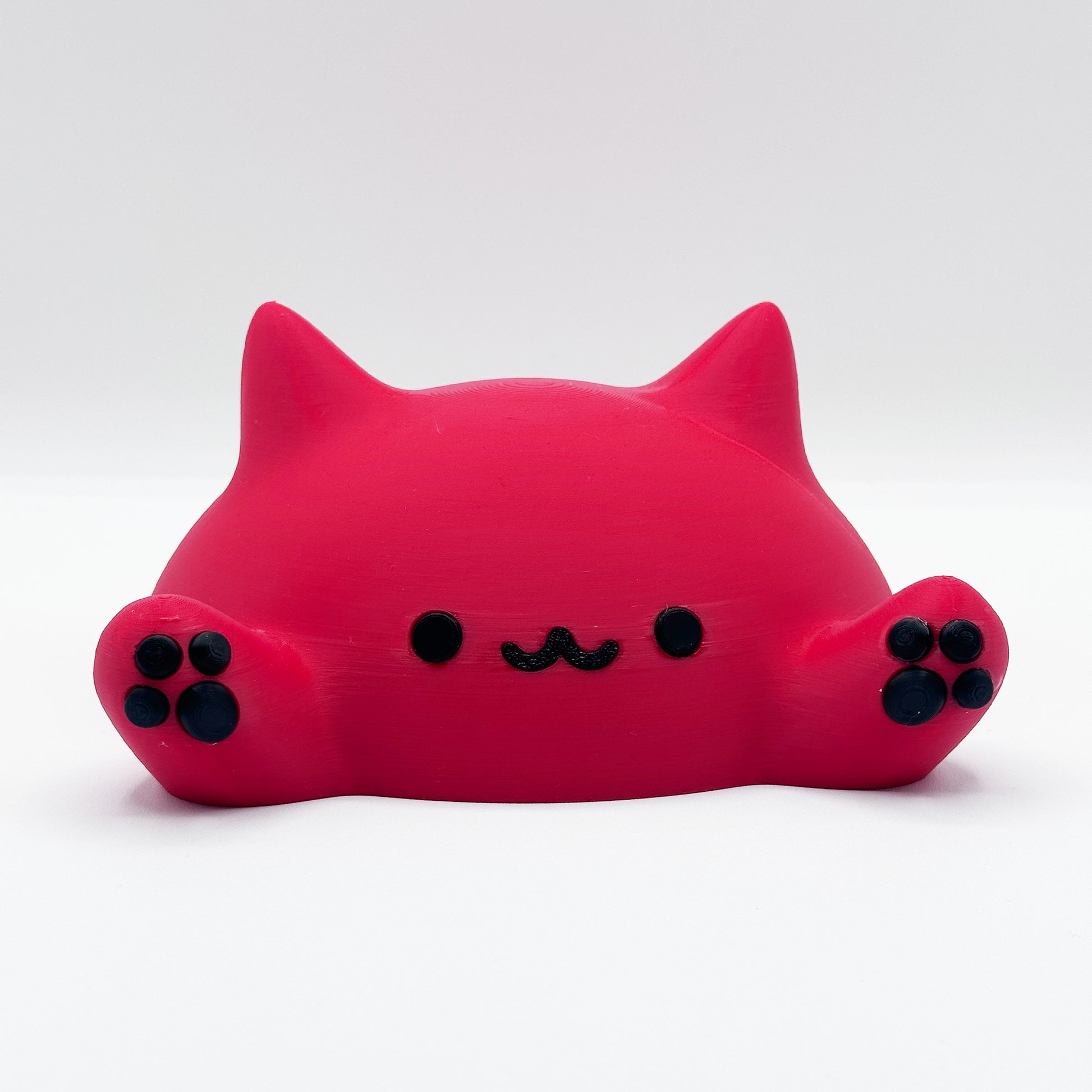 A cute, minimalistic figurine of a red cat. The cat is lying down with its head slightly raised, and has small, black, symmetrical eyes and a simple black mouth. The figurine's front paws are visible, with black paw pads. The background is plain white. This is the Blob Cat Desk Decor by Highly Obtainable.