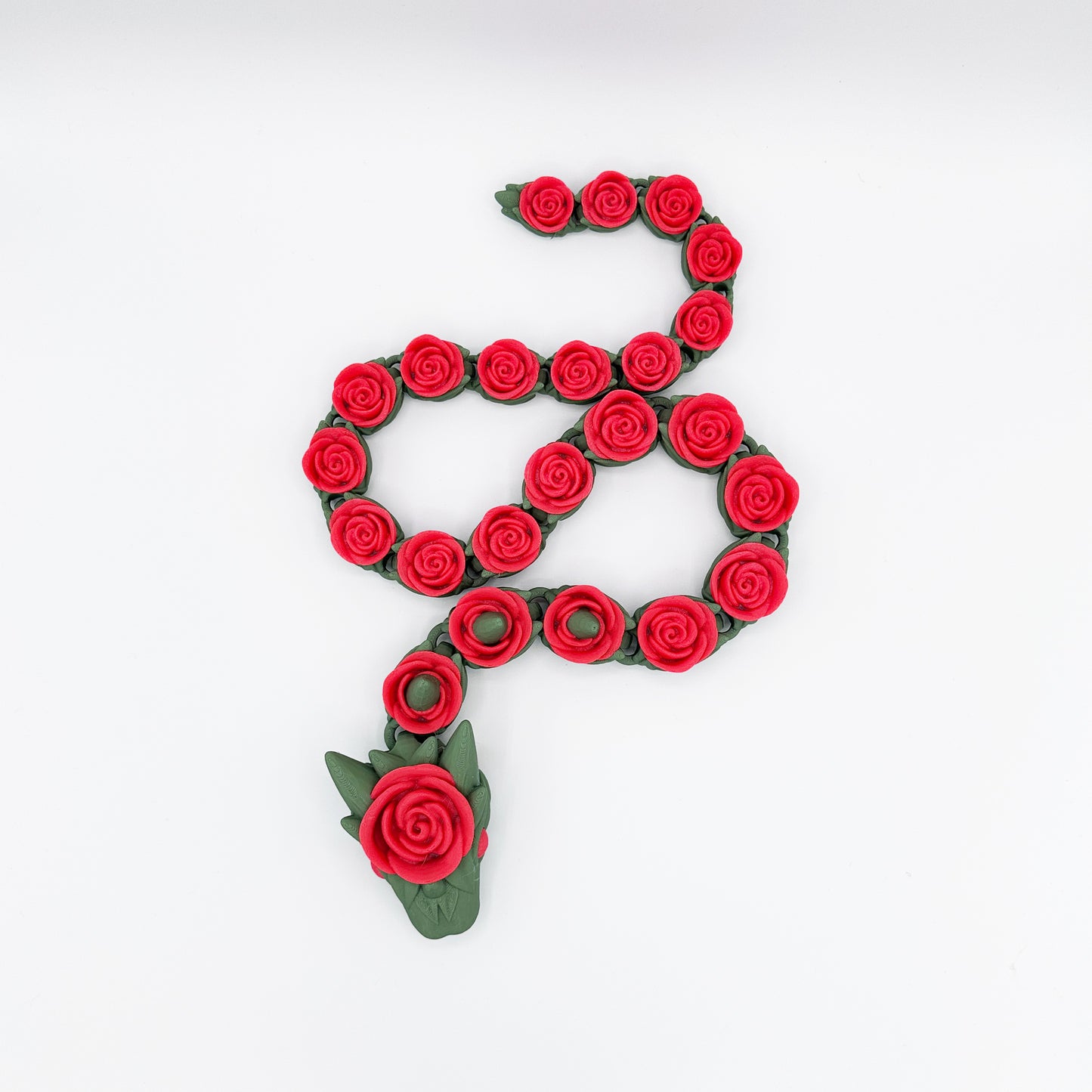 The Cinder Wing Rose Snake Dragon by Highly Obtainable is a decorative piece shaped like a snake, made entirely of red roses with green leaves. The snake's head is represented by a larger rose along with leaves and a small red detail suggesting an eye. With a product size of 27 inches, this stunning design is set against a plain white background.