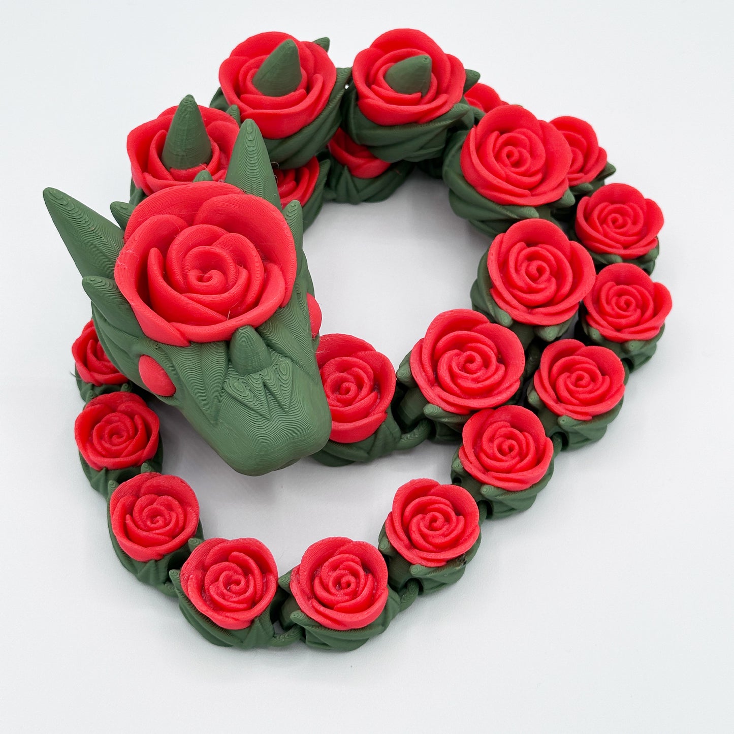 A Highly Obtainable Cinder Wing Rose Snake Dragon made of green and red clay is coiled in a circular shape. The dragon's 27-inch body and head are adorned with intricate red roses, creating a striking mix of colors and textures. The background is plain white.