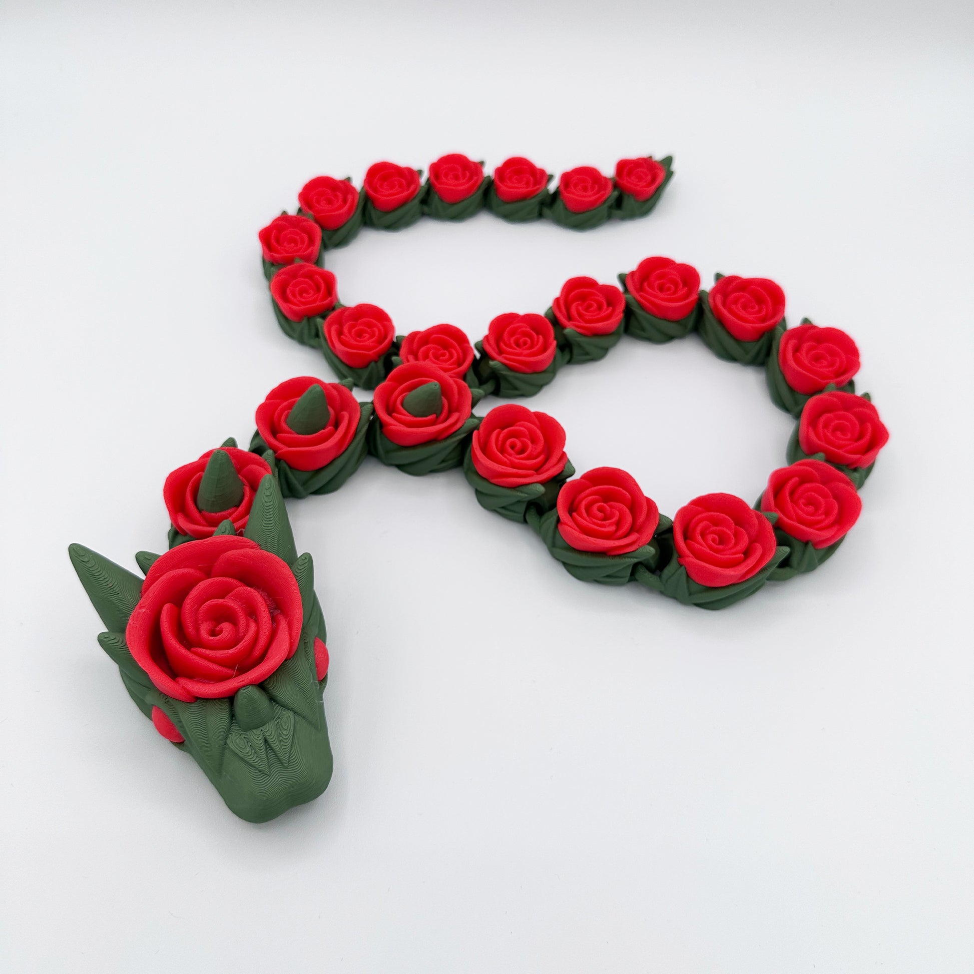 A decorative piece mimicking a dragon is made up of a chain of red roses. The 27-inch arrangement forms an 'S' shape, with the dragon's head adorned with green spikes and a large red rose at the center. The background is plain white. This is the Cinder Wing Rose Snake Dragon by Highly Obtainable.