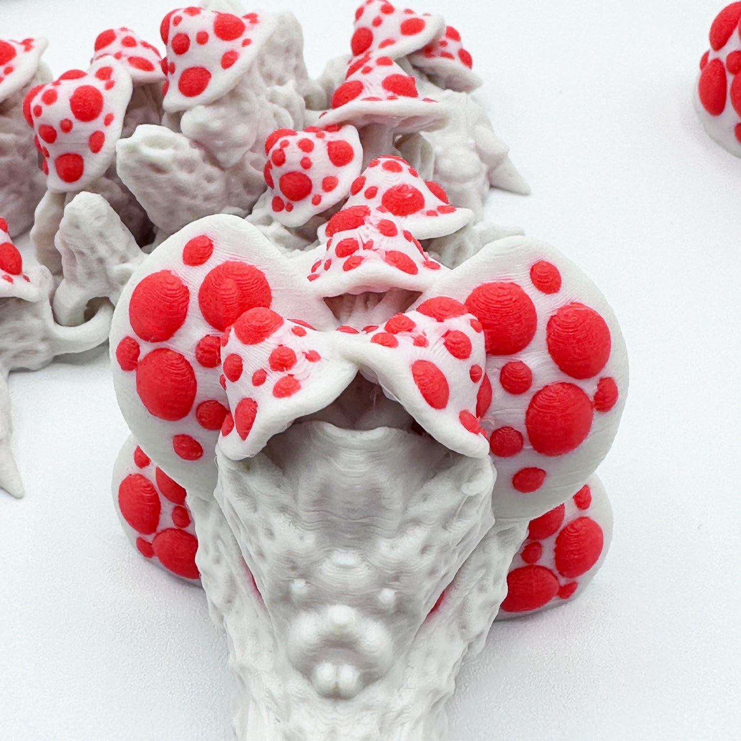 A close-up of what appears to be a group of white and red Cinder Wing Mushroom Dragons by Highly Obtainable crafted from sustainable PLA against a plain white background. The mushrooms have numerous bright red circular spots on their caps, creating a striking color contrast with the white base.