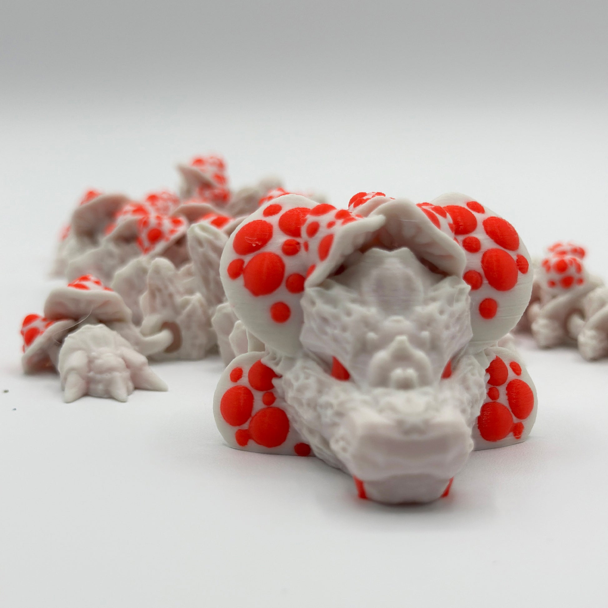 A detailed close-up of a white 3D-printed Cinder Wing Mushroom Dragon by Highly Obtainable with vibrant orange spots. The dragon appears to be lying down, with several smaller sensory figurines in the background, all crafted from sustainable PLA on a plain white surface.