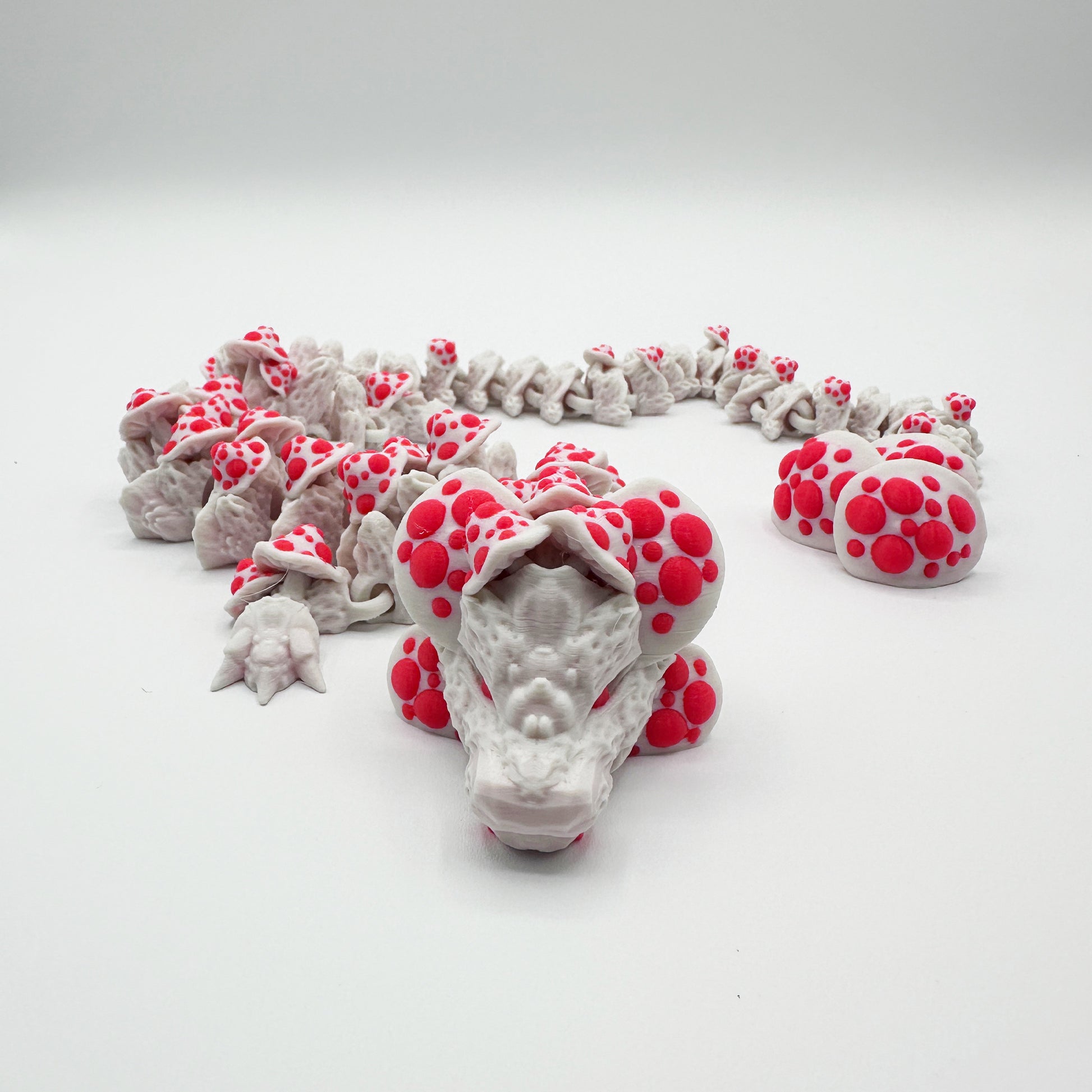 A white ceramic sculpture resembling a Highly Obtainable Cinder Wing Mushroom Dragon is shown against a plain background. The dragon's segmented, coiled body is adorned with raised, bright red circular spots, giving it a distinctive and whimsical appearance.