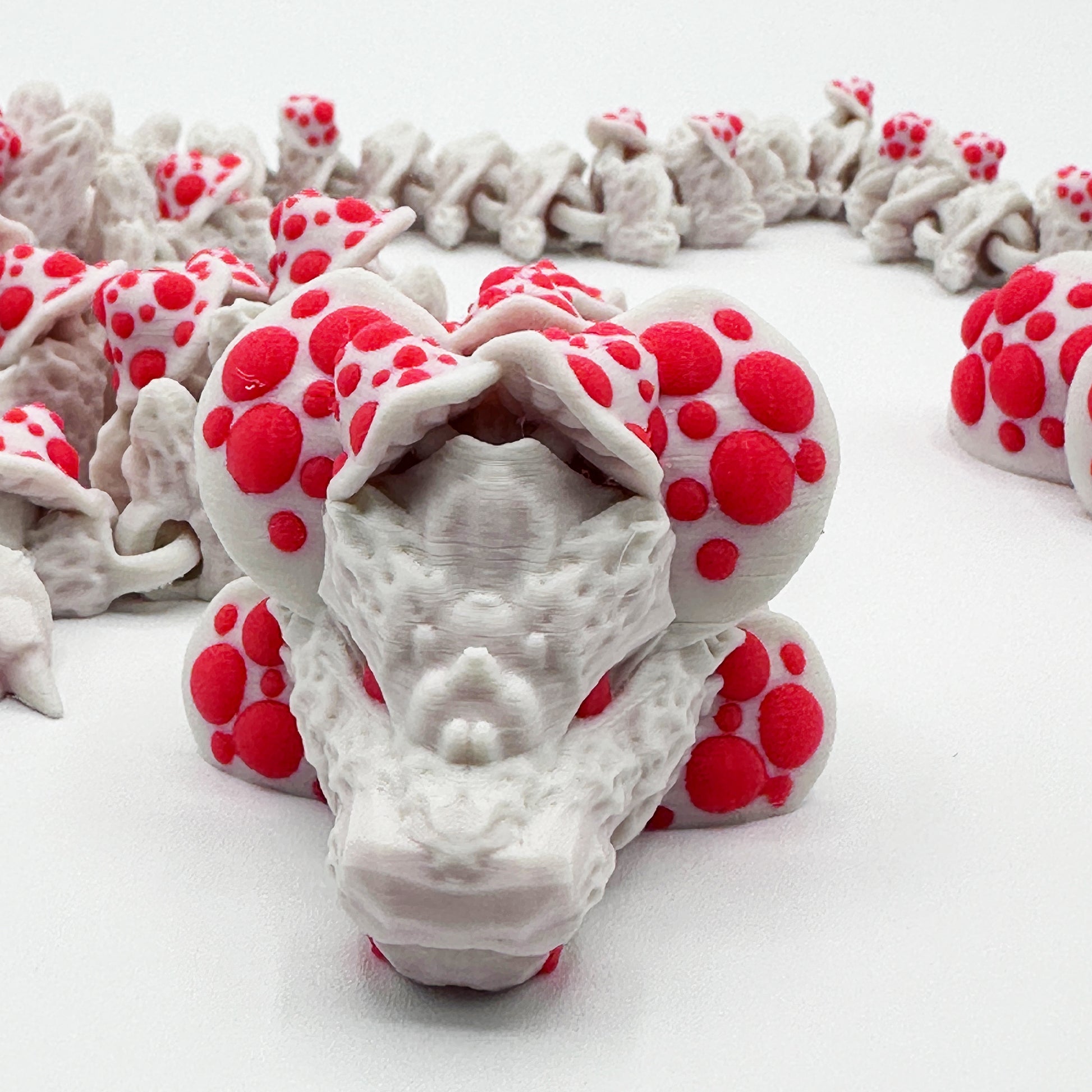 A close-up view of a **Highly Obtainable Cinder Wing Mushroom Dragon** in white with red polka dots. The dragon's intricate design includes detailed scales and patterns, creating a lifelike texture. Made from sustainable PLA, the sensory figurine is coiled with parts of its body visible in the background.